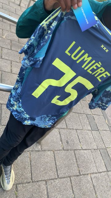 YSTHEPLUQQ community member houdt een gepersonaliseerd Lumière 25 shirt omhoog met bijbehorende YSTHEPLUQQ kaart